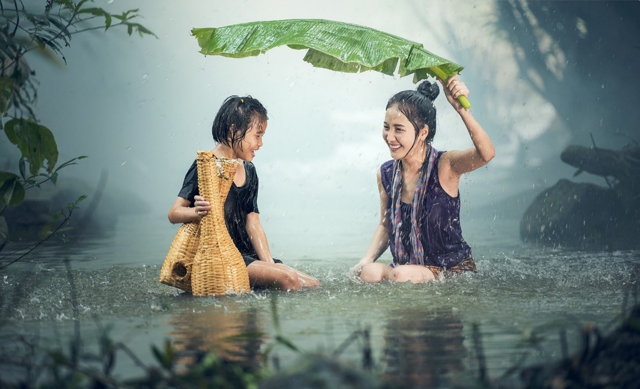 foto de uma mulher e uma menina em um lado. A mulher segura uma grande folha para proteger a menina da chuva. Ambas estão rindo